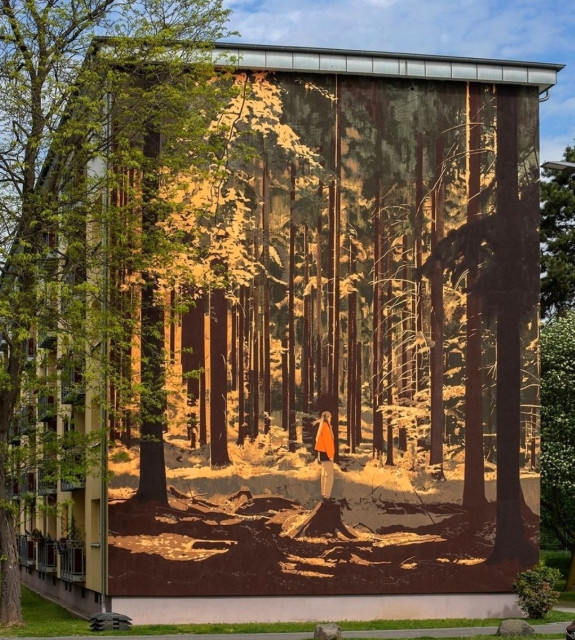 Streetartwall. A mural of a figure between tall trees has been sprayed/painted on the outside wall of a four-storey building. Several rows of tall yellow/orange trees fill the picture. Light falls on a small clearing. The lower part is in darkness. In the center, a young blonde woman stands on a tree stump, slightly elevated, looking to the right. She is wearing an orange jacket and beige trousers. Both the color composition and the dense atmosphere convey a feeling of security, but also of loneliness at the same time. (In the photo, some tall trees frame the building and merge with the wall painting)
Info: The idea for the mural came to NEAN when he heard the piece "Four" by pianist and composer Nils Frahm.
