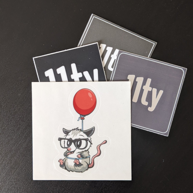 A small stack of stickers for Eleventy, three have the logo white on black, the fourth is on top, as a cute cartoon illustration of a possum wearing glasses, with a string around its waste attached to a balloon. It is considerably cute.