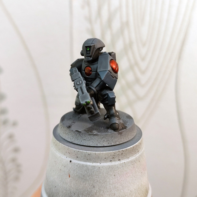 A plastic model of a kneeling T'au Fire Warrior of the Breacher kind. It is painted in a simple grey and black colour scheme, with dark blue grey and red details. 