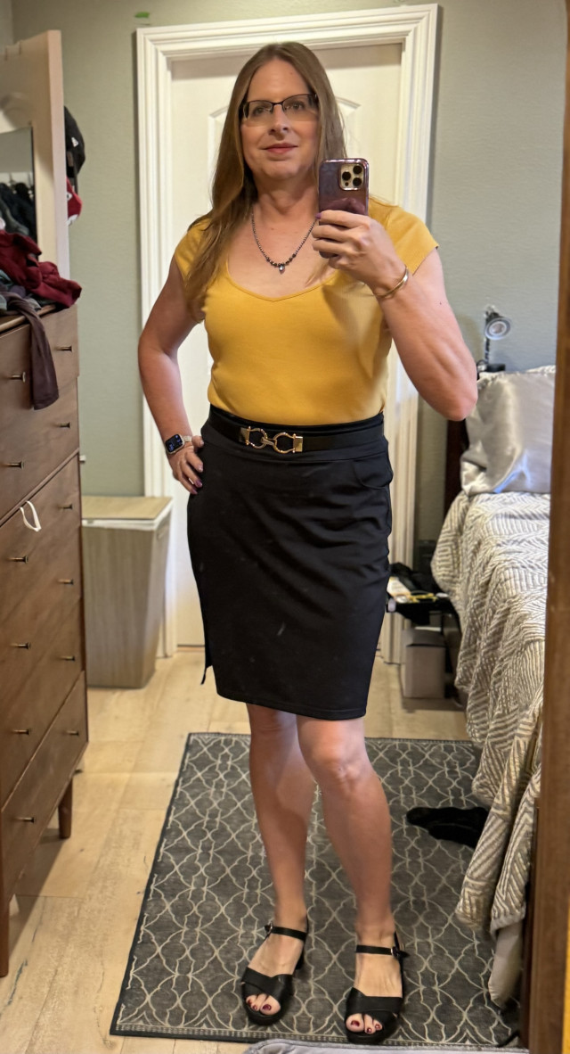 Woman taking a selfie in a mirror wearing a mustard yellow shirt, black skirt, and black heels.