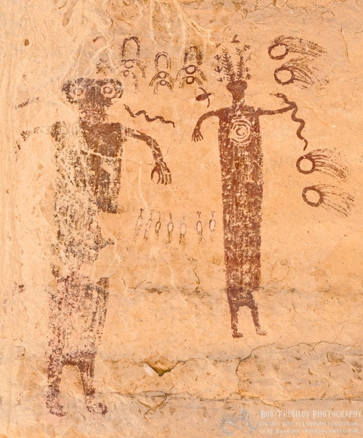 A color photo of a badly eroded pictograph painted in a red ocher. There's two anthropomorphs. The one of the right is still complete and is like a long ribbon to two short legs. The right arm is extended downward showing five figures. The left arm holds what looks like a snake at arm's length. The snake's head points toward the figure's head which has two tall hairy antenna features. The figure has an open spiral shape in its chest. The figure on the left has a long ribbon body that is mostly washed away. It is bigger than the figure on the right. Two arms extend out from the body and point downward. The head is hammerhead looking with very large eyes. A floating snake points toward the figure's left cheek. In between the two figures, at waist level, are seven egg shaped drawings that have lines extending a short distance from opposing ends. Each appears to have been painted white in the middle of the egg shape. Above and between the figure's heads are four shapes that look like two birds flying above one another with an arch shape above each set of birds. To the far right of the first figure, above and below the snake being held at arm's length, are four circles with comet like tails extending off to the right. Two are above the snake and two are below.
