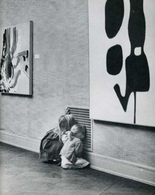 Photographie. A black and white photo of two little blonde girls in short dresses sitting on the floor in a museum and looking into a ventilation grille. Next to them hang two paintings of modern art, but the grille is more interesting.
Info: The photo comes from a publication of "The Montana Standard" from April 28, 1968.