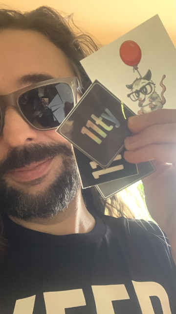 Ricky wears sunglasses and holds up several Eleventy stickers like playing cards while wearing the new Keep Building for the Web t-shirt
