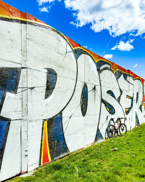 Photo of a red BMC Road machine road bike, leaning up against a wall covered in one large piece of graffiti. The graffiti consist of the word “poser” in large stylized letters. the photo is shot at an angle such that the beginning of the word is large and the end of the word is small. Above the wall is a bright blue sky with puffy clouds. Below the wall is bright green grass.