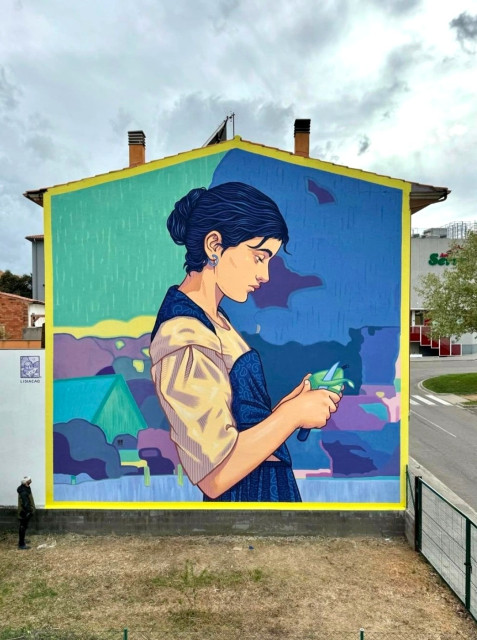 Streetartwall. A beautiful mural of a young woman peeling a pear was sprayed/painted on the outside wall of a two-story house. The young dark-haired woman wears a blue and beige dress and is depicted from the side peeling a green pear with a knife. The background is mainly blue and green and depicts a minimalist landscape. A wonderfully captured moment. (The photo shows a front garden with a meadow in front of the mural and a road next to it).
Info: The coat of arms of Calldetenes features a golden crown and three pears.