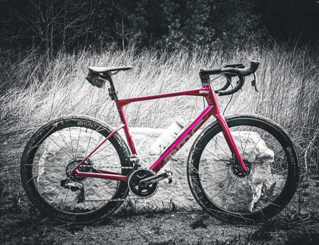 Photo of red BMC roadmachine road bike standing next to a large white boulder. behind the boulder is a patch of thick tall grass and behind the grass is a line of trees. The photo is monochrome, except for the red of the bike’s frame. 