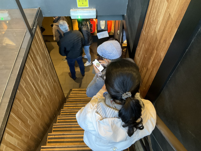 The back of 6 people queueing on a stair going down, indoors. Most surfaces are covered in wood.
Some people are holding a mobile phone, one display is visible and the viewer can guess that it is showing the menu.