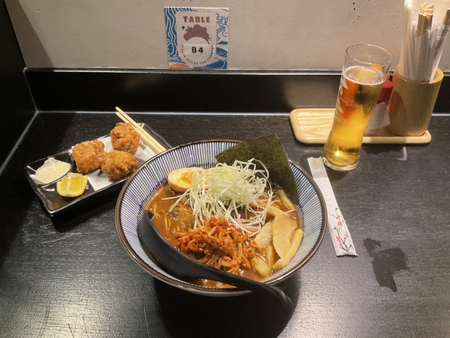Picture of a bowl of Ramen, a small plate with Karage in the back and a glass of Kirin beer next to a wooden cylinder with cutlery on the right.