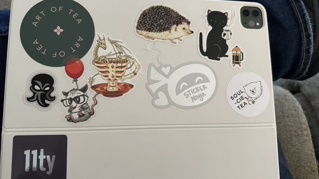 The back of an iPad Pro keyboard cover with many stickers including the 11ty possum and logo. 
