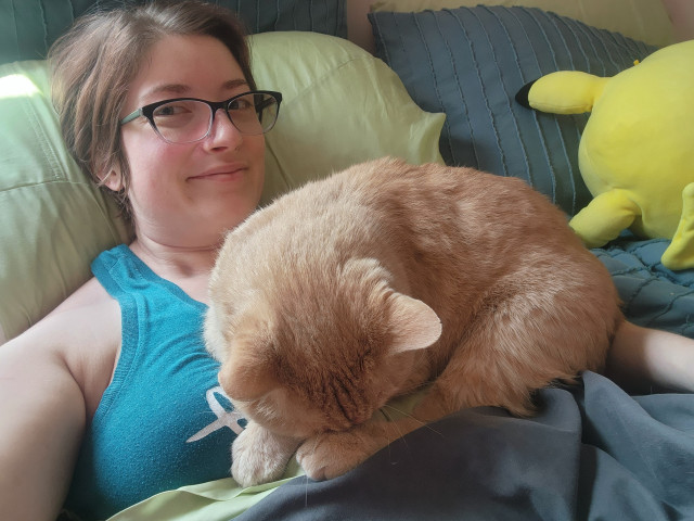 Bobby, an orange tabby Manx cat, is curled up asleep on top of his owner, Calae, who is lying in bed. Calae smiles at the camera, happy that Bobby feels secure enough to sleep on her.