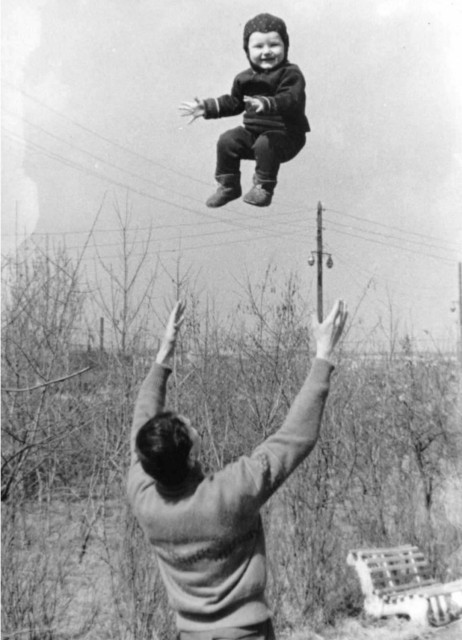 Photograph. A black and white photo of a man who has thrown his baby into the air. The photograph shows the moment when the father, standing with his back to the photographer, raises his arms and the child floats. The small child, dressed in winter clothes, floats in the air about 1 meter above him (in a kind of sitting position with outstretched arms) and laughs towards the camera. (The background of the photo consists of thin leafless trees and a white bench at the bottom right of the picture). 
Info: French photographer René Maltête (1930-2000) was a passionate street photographer. His photos are simply amazing, and what is even more impressive is the sheer number of fleeting moments he was able to capture. He was characterized by a mischievous sense of humour, nonchalance, gentle irony and sometimes seriousness.