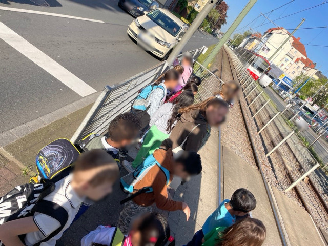 About ten children and three adults squeeze onto a traffic island between a car lane and the tracks of the light rail.