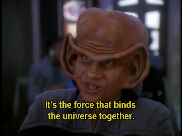 DS9 scene. Nog is pictured up close and in dialogue, I think in a cafatorium or bar. He's a Ferengi, and has giant ear lobes that run from the bottom of his ears all the up and around and down the forehead and connect at the bridge of the nose.  He's also bald and has a high rise head, kind of shaped like a beluga whale. He's explaining something here with enthusiasm. Closed caption reads, "It's the force that binds the universe together."