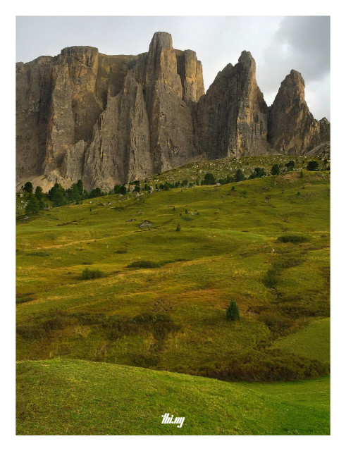 A pretty empty and slightly bumpy high alpine mountain meadow with low grass in different shades/patches of greens and browns leading directly towards the base of the 400-500m tall Sella towers, craggy, sharp, tooth-like, vertical rock faces of characteristic dolomite, rising up to ~2600m. Gray sky. The entire scene almost feels like a minimalist landscape painting.