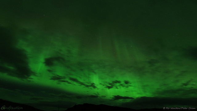 A photo of the sky at night. Stars can be seen behind columns of green aurora, seemingly stretching up to infinity. It looks like a very fine gauze curtain lit from below by soft green light, moving gently in a draught of air. It's behind a layer of low cloud so that part of its beauty is obscured. At the very bottom of the shot is the dark silhouette of mountains, suggesting scale.