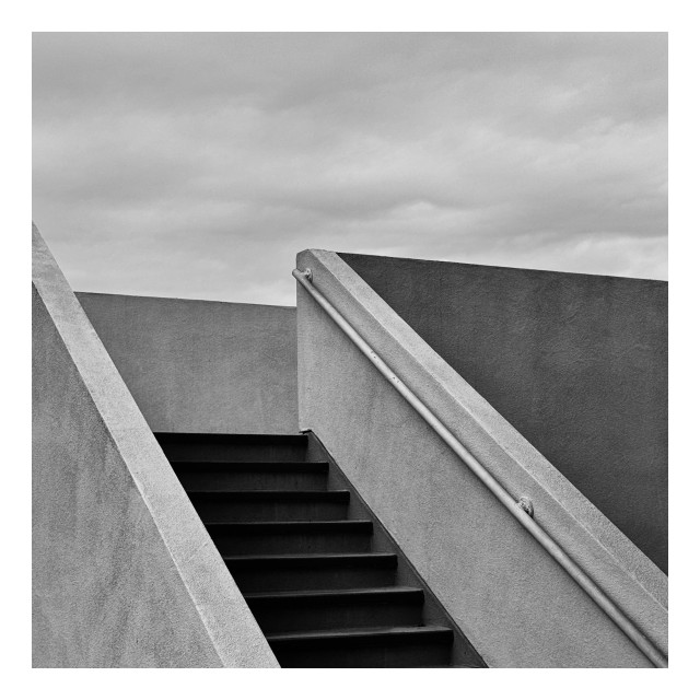 This image captures an architectural detail in black and white, focusing on a set of concrete stairs ascending diagonally across the frame. The stairs are bordered by thick, smooth concrete walls with angular, clean lines that emphasize a modernist aesthetic. A handrail runs alongside the stairs, enhancing the linear perspective. The background shows a flat, overcast sky which provides a neutral backdrop that complements the stark geometry of the structure. The composition highlights the interplay of light and shadow on the surfaces, giving a sense of depth and solidity to the concrete elements.