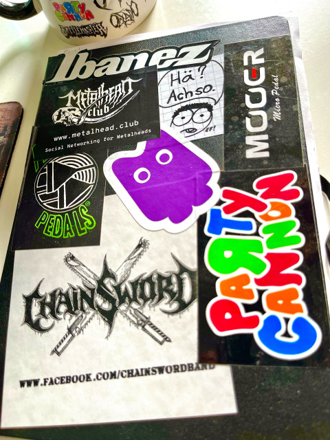 A notebook with stickers on it, from metalhead.club, Mooet (pedal company), Ibanez, Chainsaw and Party Cannon (bands), and more.