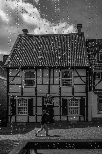 Eine junge Frau geht in der Altstadt von Essen Kettwig an einem Fachwerkhaus entlang. Sie wirft einen Schatten auf das Kopfsteinpflaster. Im Vordergrund sind Wassertropfen zu sehen, die wie ein Vorhang wirken.
Das Foto ist in schwarzweiss. 