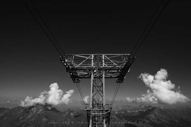 Contrasting black and white photograph of a cable car mast. Two black cables run from the centre of the frame and disappear into the blackness of the sky above. The mountains can be seen on the horizon and above them, on either side of the mast, two white clouds stand out against the grey mountains.