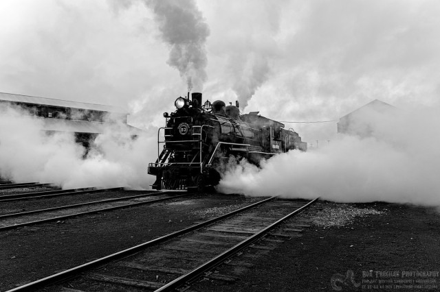 A modern black and white photo of a old steam engine. It's a grey rainy day. The steam engine is blowing out the side cylinders so there's lots of steam spraying out both sides in the front. There's steam coming out of the whistle and a grey smoke coming from the smokestack. 