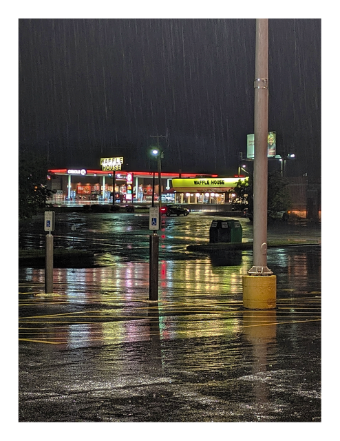 night. lit by streetlights. in the foreground an empty, wet supermarket parking lot with light pole. in the mid-distance, a waffle house with one car in front, headlights on. across the highway, a circle k store with lighted, red fuel canopy in front. 