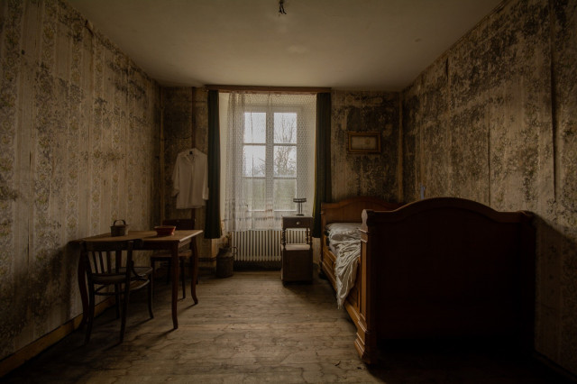 Ein Schlafzimmer in einem verlassenen Haus. Links neben dem Fenster hängt ein einzelnes weißes Hemd. 