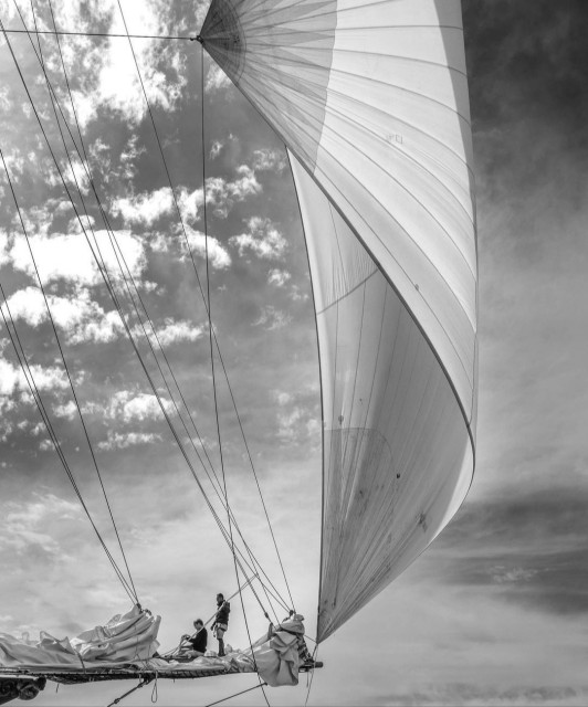 Photography. A black and white photo of a sailing boat with an upright, hoisted sail. The wind is blowing into the sail. On the right is a gray sky with a few gray veils. On the left is a horizontal mast with two boatmen on it. They are standing in the rigging and arranging the ropes. A sky with many white clouds can be seen above them. A beautifully arranged sailing photo with an interesting division of light and sky.