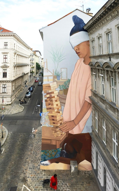 Streetartwall. On a narrow exterior wall of a long, three-story building, a beautiful mural of a blindfolded young woman has been spray-painted/painted. She has dark hair and is wearing a pink T-shirt and brown trousers. A black blindfold is tied over her face. In her hands she is holding a Jenga tower made of small wooden blocks, which she is trying to place on a table. It already seems to be falling. A room can be seen in the background. (The photo shows a woman in a red coat with a trolley case in front of the huge mural and next to it a view of the street with its old canyons of houses)
Info: The Artist: "Playing Jenga with your eyes closed, recon only on your sense of touch, could be quite challenging. Mural is picturing the moment before falling Jenga activates the sense of hearing, the moment in which it is possible for an expected surprise to happen.”