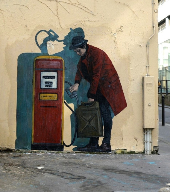 Streetartwall. A self-designed, life-size paste-up showing a young man at a petrol pump was glued to a yellowish exterior wall. The dark-haired man in a long, wine-red coat, hat, jeans and black shoes is standing next to an old, red petrol pump with a petrol can and is pouring petrol into it. However, his shadow behind him shows that he is holding the tap in his mouth and drinking. Title: "Thirst"
Info: The artist Charles Leval, alias Levalet, is now exhibited in museums. He stands out from the usual forms of street art, as his scenes, lovingly drawn with ink on paper, show figures in their (often tragic-comic) everyday lives. They seem like repressed, unconscious feelings and experiences. They often fill long walls and are even told as serial stories.