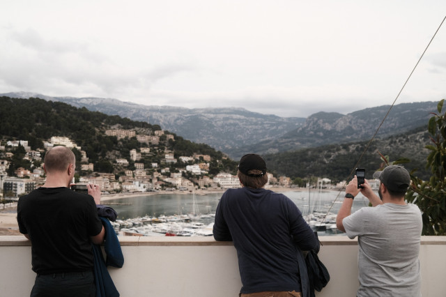 Three men taking photos of a coastal scene with mountains, buildings, and a marina with boats.