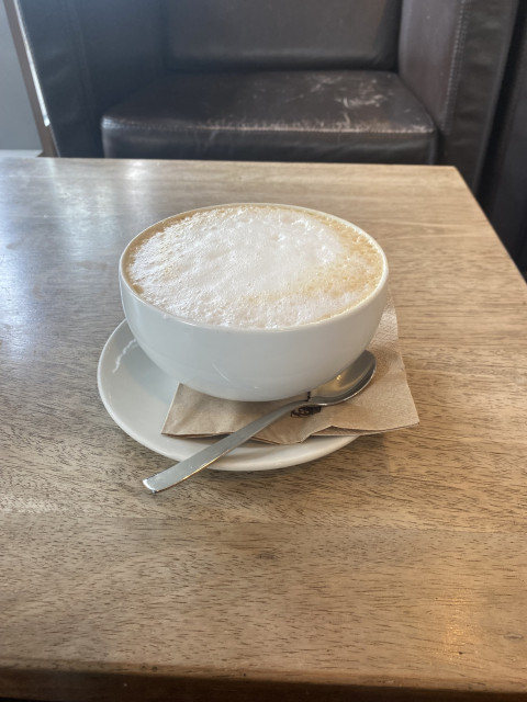 Ein Photo einer Tasse Milchkaffee auf einem Tisch, im Hintergrund ein etwas angewetzter schwarzer Ledersessel.