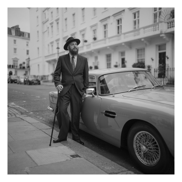 Portrait of a man dressed in vintage clothing with a cane, standing near a classic Aston Martin.