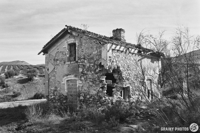 A black-and-white film photo of an abandoned level-crossing keeper's cottage by the via Verde. On one corner the wall has crumbled and there is a gaping hole.