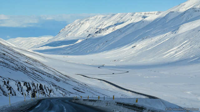 A photo of a snowy landscape under a cloudy blue sky. A valley is in front of the viewpoint, with tall mountains on either side covered in ice and snow. Snaking through the centre of the valley is the black line of a tarmacked road, some traffic can be seen on it. The viewer is on that road, high above the valley and about to drive down into it. On either side on the valley floor is more snow and ice. There are cloud shadows across the snowfield, and the light is from behind the viewer.