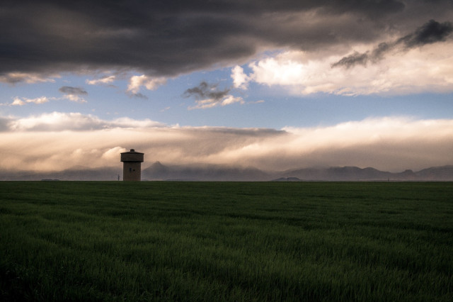 Fotografía con un cielo lleno de nubes de tormenta con algunas montañas al fondo y un prado y una torre de agua en primer plano.
