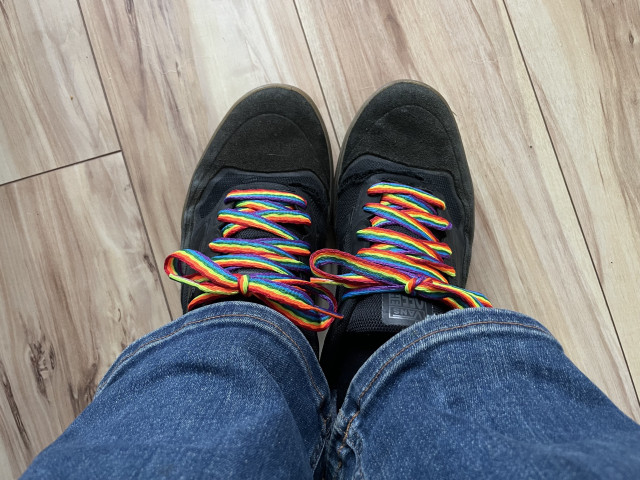 Photo of black Vans shoes with rainbow laces. Person is wearing jeans and the floor is light wood laminate.