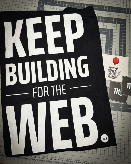 A black t-shirt with the words "Keep Building for the Web" and an 11ty icon in white, next to it are three stickers, one 11ty possum mascot with a red baloon, and two 11ty logos on black background. The items are all on top of a grey cutting mat of the craft table.