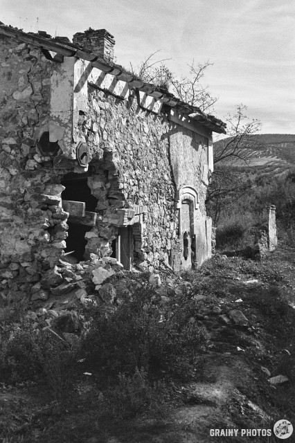 A black-and-white film photo of an abandoned level-crossing keeper's cottage by the via Verde. The wall has crumbled on one corner and there is a gaping hole.