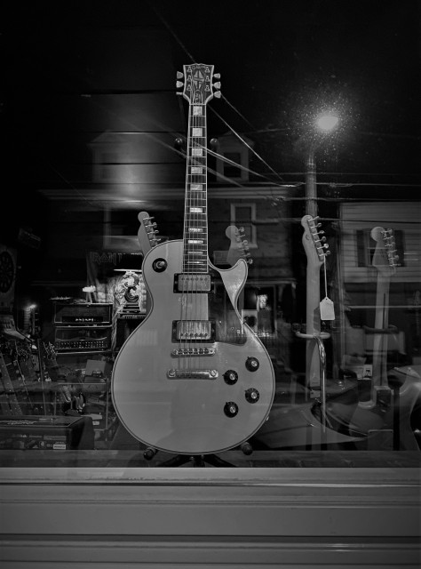 Black and white photo of the window of a vintage guitar shop taken in the dark, wee hours. The focus is on a Gibson electric guitar in the middle of the frame. In the background is a muddled mix of items caused by the reflection in the glass. There are other guitars in floor stands, amplifiers, and an Iron Maiden record album. But also in the shot is an automobile parked in front of the window, houses across the street, a street lamp and electrical wires all caused by the reflection.