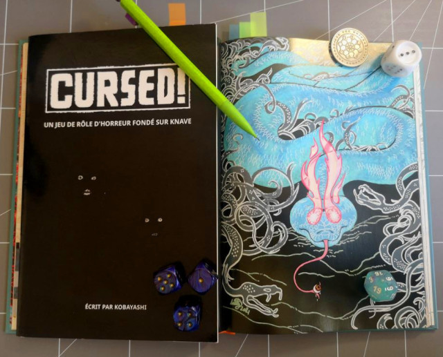 Two tabletop role-playing books. The first one is the game "Cursed", by Kobayashi. The second one is "A Folklore Bestiary" by The Merry Mushmen, opened on the page depicting the Boitata, a blue snake with flaming eyes about to devour a human eye currently sitting on the tip of its tongue.
The second book has little bookmarks to mark the creatures from Brazilian folklore.
Dice, a coin and a pencil sit on top of the books.