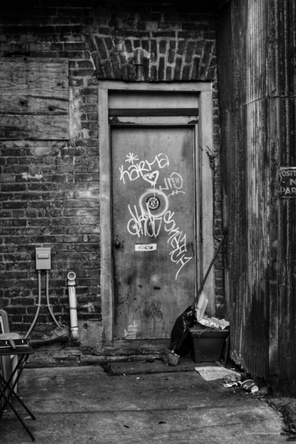 A back door in an alley is seen in this black and white image. Graffiti covers the door and trash on the ground makes up this moody photo.
