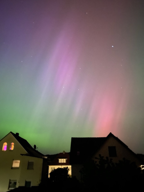 Aurora borealis in pink and green with an upwards striped pattern. A few houses are seen against the colorful night sky. 
