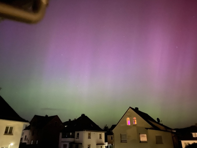 Aurora borealis in pink and green looking somewhat hazy. A few houses are seen against the colorful night sky. 