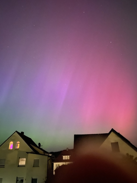 Aurora borealis in bright pink and pale green with a slight upwards striped pattern. A few houses are seen against the colorful night sky. 
