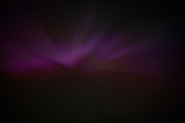A dark photo looking upwards to the night sky, showing the Aurora Borealis.
There is one predominant magenta colour, with wavy patterns running through it.