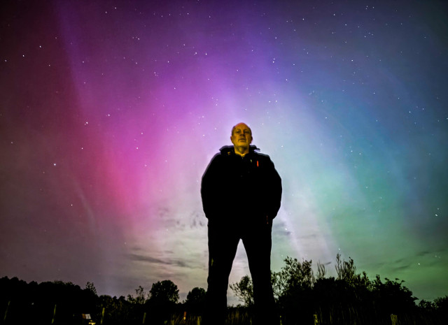 A long exposure selfie taken from ground level with an iPhone 15, showing me dressed in back illuminated by some nearby street lights, against a bright purple, red, blue, and green aurora. There are also some trees behind me.