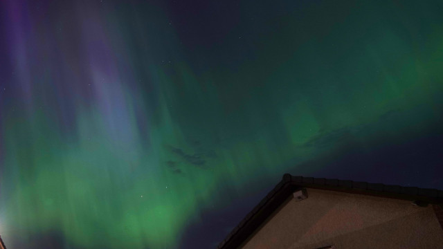 A photo of the night sky above a bungalow roof. Vivid green streaks shoot down in the sky, with blue and purple among them.