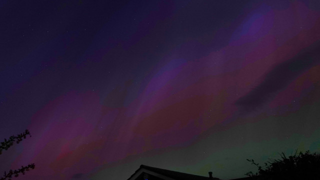 A photo of the night sky above a bungalow with blue/purple at the top left, a pink/red/orange band across the middle, and a green glow at the bottom