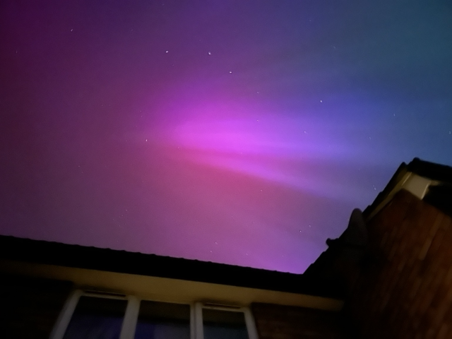 Picture of the Aurora borealis, seen over the roof of a brick house. There are bright purple and blue streaks from left to right