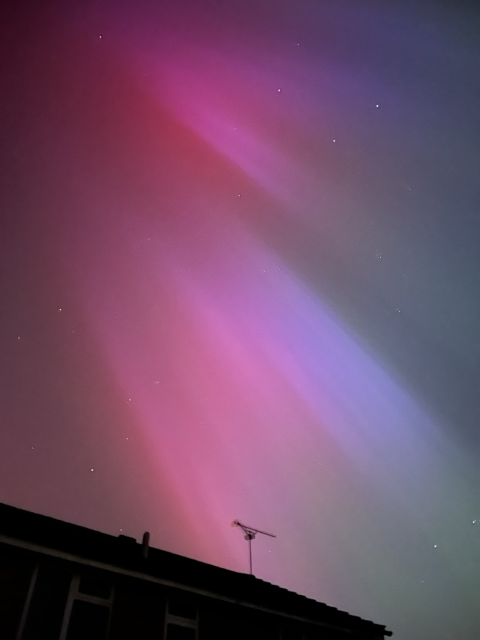 Picture of an Aurora borealis, with some stars just visible. Pink, purple, and green diagonal, almost vertical streaks. The top of a roof is just visible at the bottom of the picture as a dark silhouette.